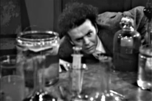 Screenshot from the film Maniac (1934), public domain. A character looks at glassware on a countertop.