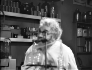 Screenshot from the public domain film Maniac (1934). The camera is out of focus and showing Horace B. Carpenter as the character "Dr. Meirschultz" behind lab equipment.