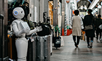 Photograph of a white and silver robot holding a tablet in front of a luggage store. In the background, in the hallway, two people are walking with their backs to the camera.