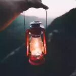 A photograph of a hand holding a lit flashlight against a forest backdrop in daylight.