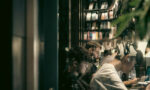 A photograph of researchers reading on laptops through the glass of a library, with books in the background.