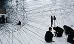 Fotografía de una obra de arte compuesta por dos estructuras en forma de globo hechas y levantadas por cables de acero. En primer plano, tres personas están arrodilladas mirando hacia donde uno de los cables está sujeto al suelo justo al lado de uno de los globos. Al fondo, junto al otro globo, hay dos personas de pie hablando.