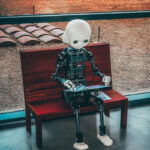 Humanoid robot with a white head sits on a wooden bench by a large window, reading a brochure or leaflet.