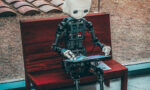 Humanoid robot with a white head sits on a wooden bench by a large window, reading a brochure or leaflet.