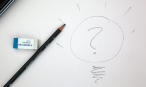Fotografia de uma folha de papel em que está rascunhada em lápis uma lâmpada com uma interrogação dentro. Ao lado esquerdo do desenho está um lápis e uma borracha.