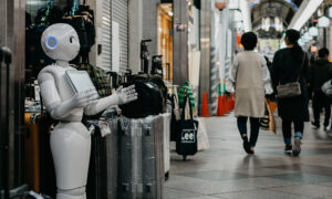 Fotografia de um robô branco e prateado segurando um tablet na frente de uma loja de malas de viagem. Ao fundo, no corredor, duas pessoas estão caminhando de costas para a câmera.