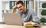 Fotografia de um homem usando óculos e fones de ouvido sorrindo enquanto olha para a tela de um laptop. Ele está sentado em uma mesa com um livro, cadernos e uma caneta. Ao fundo, há prateleiras com pastas e vasos com plantas.