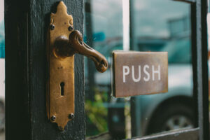 Fotografia de uma porta de madeira pintada de preto com vidro e uma maçaneta alavanca que está sendo virada, de forma a abrir a porta. Ao lado da maçaneta há uma placa onde se lê "Push".